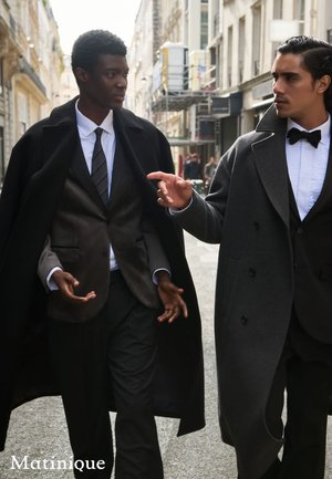 Two men in tailored suits walk on a city street. One wears a black cape, the other a grey overcoat, both paired with formal attire.