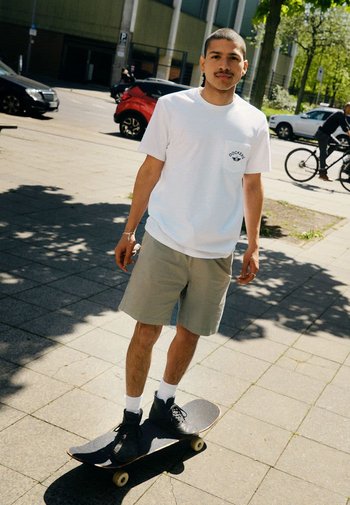 Planche à roulettes avec un plateau en bois, un grip noir et des roues blanches. La personne porte un t-shirt blanc, un short kaki et des baskets noires.
