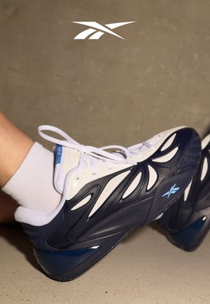 Persona che indossa scarpe da ginnastica bianche e blu navy con lacci bianchi e calzini alla caviglia bianchi, su un pavimento e una parete beige.