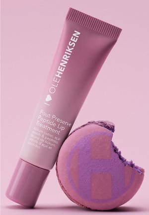 Tube de traitement pour les lèvres Ole Henriksen Pout Preserve Peptide à côté d'un macaron violet partiellement mangé sur fond rose.