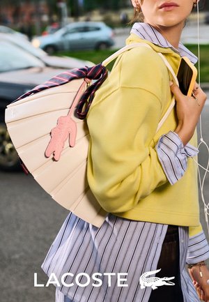 Mujer con camisa de rayas en capas y suéter amarillo lleva un bolso beige en forma de abanico con un charm de cocodrilo rosa y un teléfono en la calle de la ciudad.