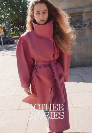 Jonge vrouw loopt buiten met een lange mauve trenchcoat met een hoge kraag en een gestrikte taille op een zonnige dag.