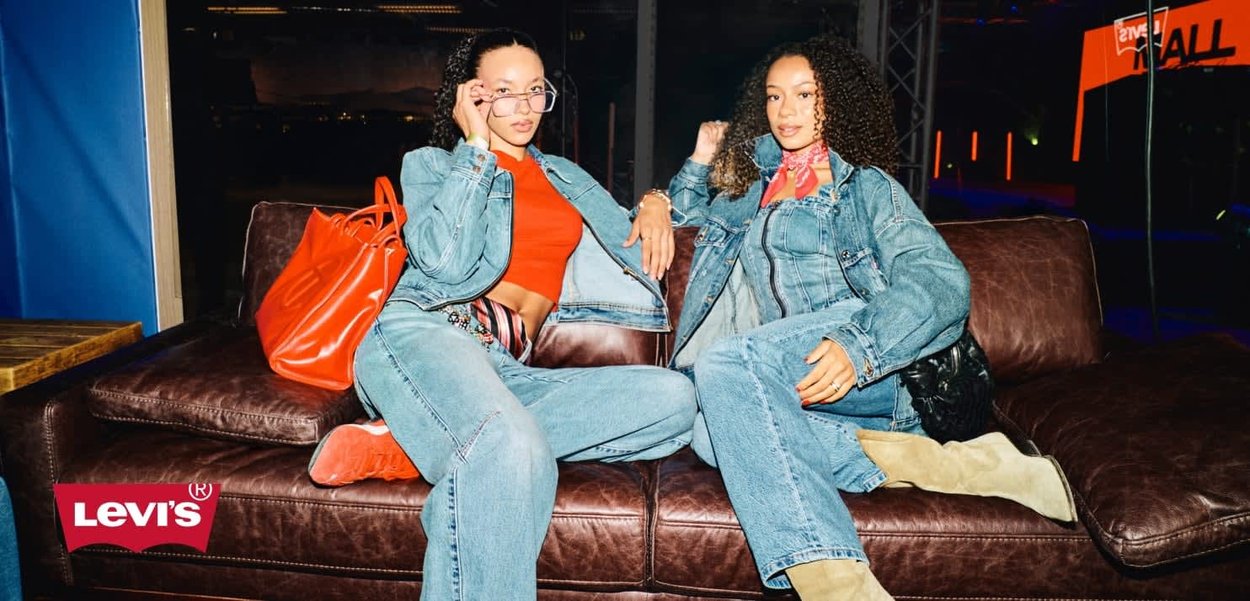 Two models wearing denim jackets and jeans, a red top, and beige boots, seated on a brown leather sofa. An orange handbag is nearby.