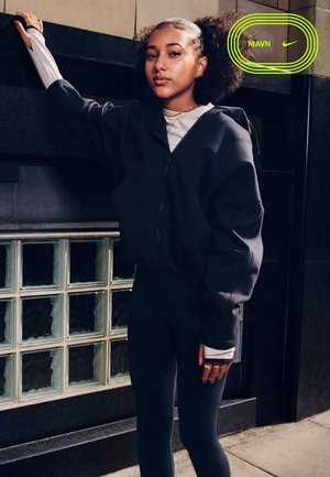 Junge Frau mit lockigem Pferdeschwanz lehnt an einer Eckwand, trägt eine schwarze Jacke mit Reißverschluss und Leggings und steht neben einem Glasbausteinfenster.