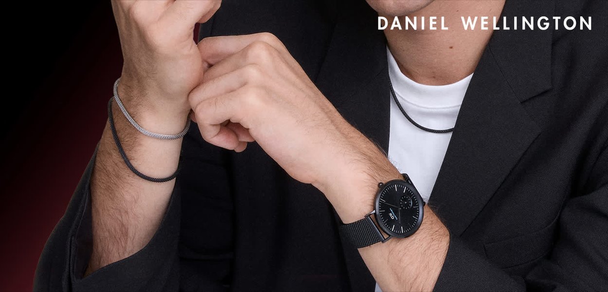 Zegarek z czarnym paskiem z siateczki z srebrnymi akcentami, zestawiony z czarnymi i srebrnymi teksturowanymi bransoletkami. Noszony na ciemnej kurtce, pod którą znajduje się biała koszula.