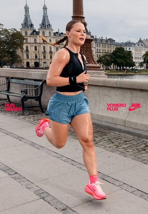 Coureur en haut sans manches noir et shorts bleu clair, portant des chaussures Nike roses. La sueur brille sur la peau ; paysage urbain en arrière-plan.
