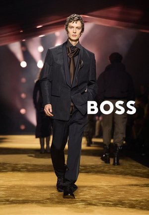 Mannequin masculin défile en portant un costume noir avec une large écharpe nouée sous un projecteur, silhouettes floues et logo BOSS en arrière-plan.