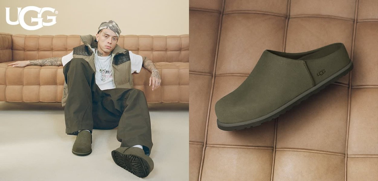 Homme portant des chaussures UGG olive vert à enfiler, assis par terre devant un canapé en cuir beige, assorties à une tenue décontractée.