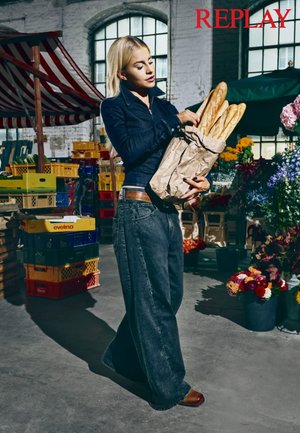 En kvinne holder en papirpose med baguetter, iført en mørk denimjakke og løstsittende blå jeans, i en markedsetting med fargerike kasser.