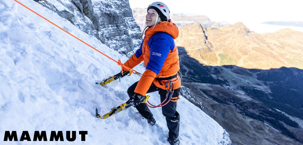 Climber in orange and blue jacket ascending a snowy mountain slope with ice axes, wearing helmet and safety rope harness.
