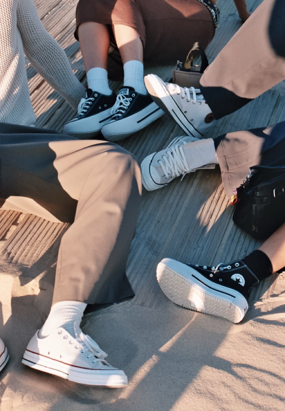 Multiple pairs of sneakers on a sandy surface: black high-tops, white low-tops, textured soles, contrasting laces, various sock types.