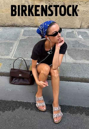 Femme portant des sandales Birkenstock imprimé léopard, chemise noire, foulard bleu et lunettes de soleil, assise sur le trottoir avec un sac à main marron à côté d'elle.