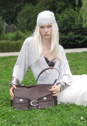 Hellbraune Lederhandtasche mit silberner Hardware, zwei Fronttaschen und einem geschwungenen Tragegurt. Ein Model in einem grauen Wickeloberteil und einem weißen Rock sitzt auf Gras.