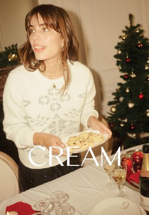 Leuchtend cremefarbener Pullover mit silbernem Schneeflockenmuster, der einen weißen Teller mit Schokoladenkeksen hält, vor einem festlich gedeckten Tisch und Weihnachtsbaum.