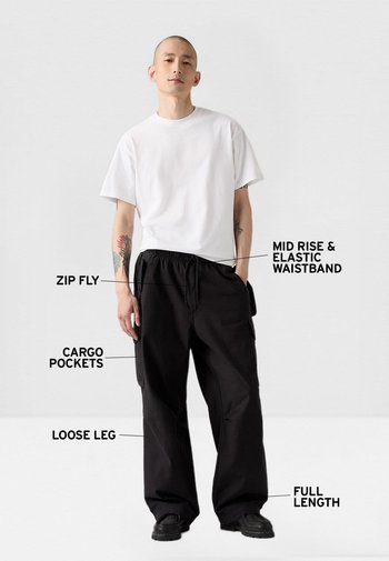 Pantalones negros de pierna ancha con cintura elástica de tiro medio, bragueta con cremallera y bolsillos cargo. Diseño de longitud completa. Combinados con una camiseta blanca lisa.