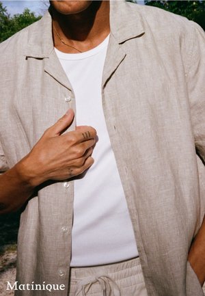 Mann trägt beiges Leinenhemd über weißem T-Shirt mit passenden beigen Kordelhosen, steht draußen mit der Hand nahe der Brust.