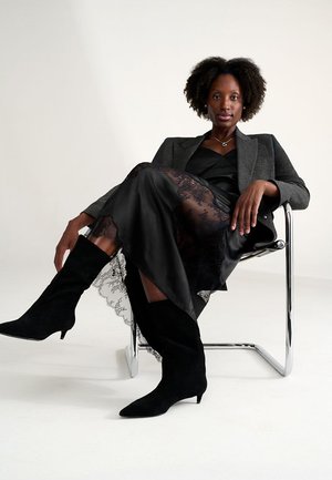 Una modelo se sienta en una silla de cromo, vistiendo un blazer gris sobre un vestido negro de satén con detalles de encaje y botas altas de gamuza negras.