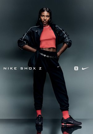 Calzado Nike Shox Z en negro, con acentos rojos; el modelo lleva una chaqueta negra sobre un top corto rojo y joggers negros. Detalle de cinturón de cadena plateada.