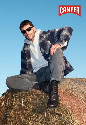 Un uomo con occhiali da sole, giacca scozzese, felpa grigia e stivali neri è seduto su un rotolo di fieno sotto un cielo blu limpido con il logo Camper sopra.