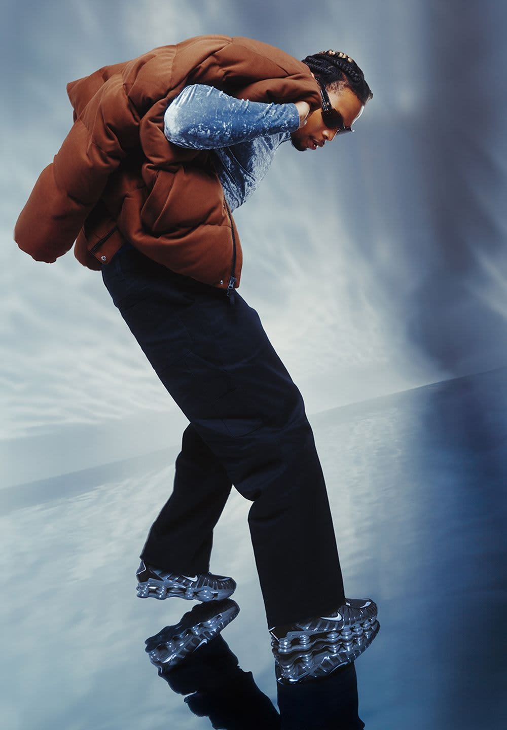 Brown puffer jacket, blue textured shirt, black pants, and metallic sneakers with a wavy design. Background features soft light reflections.