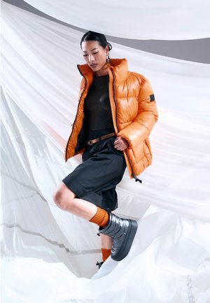 Orangene Daunenjacke mit hochgeschlossenem Kragen, schwarze Shorts und knielange schwarze Stiefel. Das Model trägt orangefarbene gerippte Socken und steht auf weißem Stoff.