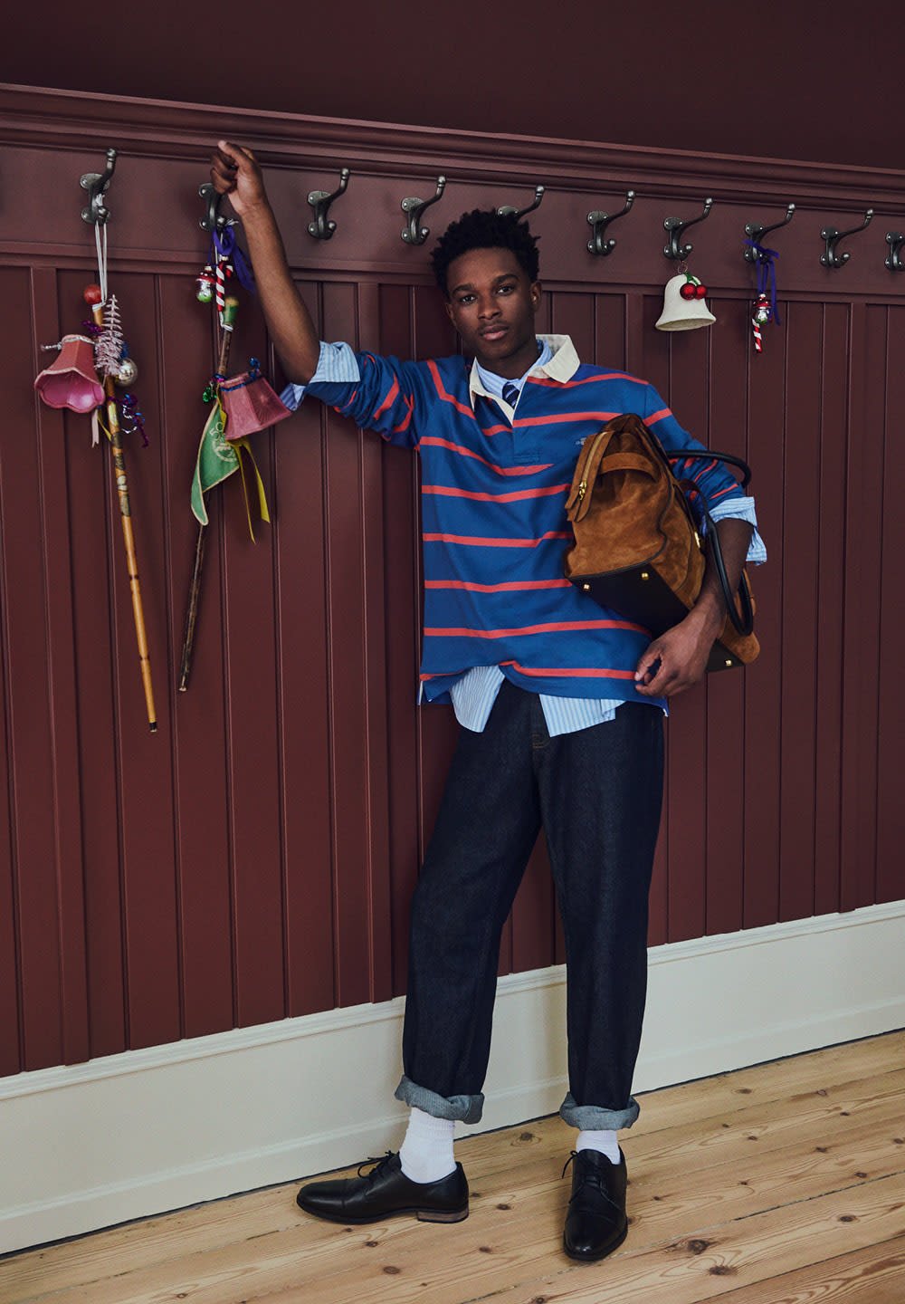 Siniraidallinen sininen ja punainen rugby-paita vaaleansinisen paidan päällä, tummat farkut, mustat kengät ja ruskea nahkalaukku, nojaamassa koukkuja vasten.