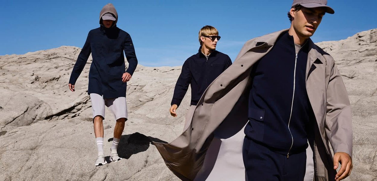 Drie mannen dragen stijlvolle jassen en zonnebrillen terwijl ze over rotsachtig terrein lopen onder een heldere blauwe lucht.
