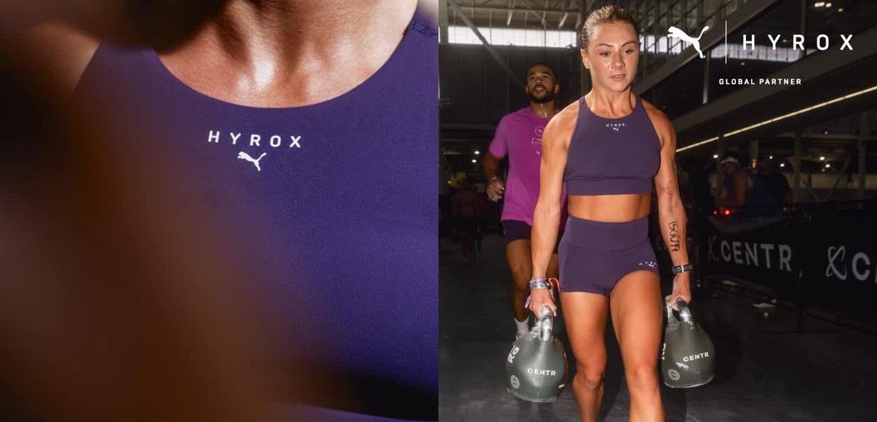 Athlète féminine en tenue d'entraînement violette portant deux kettlebells lors d'une compétition de fitness en intérieur, avec un coureur masculin derrière elle.