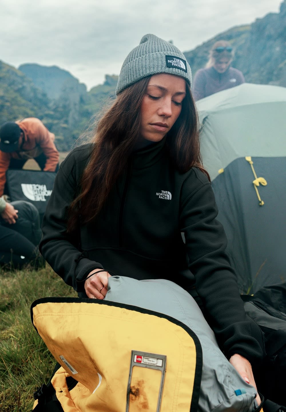 Giacca in fleece nera con colletto alto, berretto lavorato a maglia grigio e zaino giallo, parzialmente aperto, che rivela contenuti grigi all'interno.
