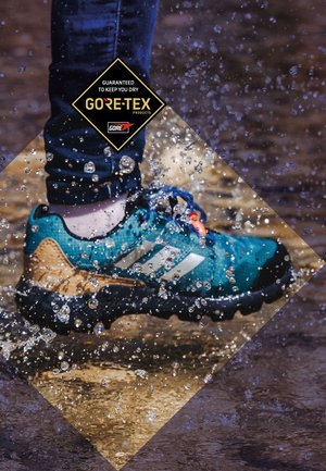 Chaussure imperméable bleue et beige avec une surface texturée, étiquette GORE-TEX et trous de drainage bien visibles, éclaboussant à travers l'eau.
