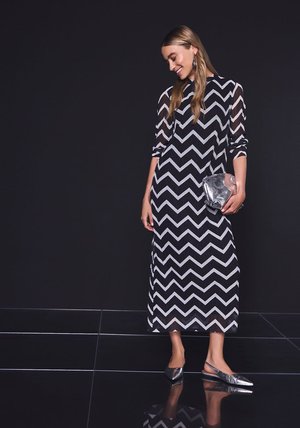 Robe maxi à motif chevron noir et blanc avec manches longues transparentes. Le mannequin tient une pochette argentée et porte des ballerines métalliques sans lacets.
