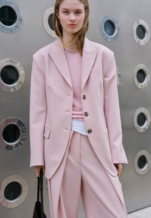 Młoda kobieta ubrana w duży, jasnoróżowy garnitur i sweter, stojąca przy metalowej ścianie z okrągłymi oknami, trzyma czarną torbę.