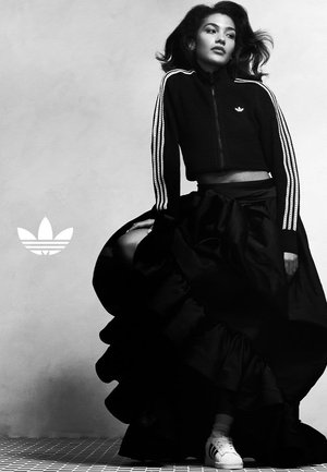 Frau in einer schwarzen Adidas-Trainingsjacke, einem fließenden schwarzen Rock und weißen Sneakers posiert vor einem schlichten Hintergrund, das Adidas-Logo ist sichtbar.