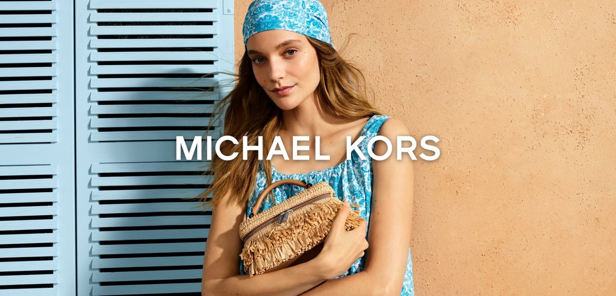 Mujer con vestido azul estampado y pañuelo a juego en la cabeza sostiene un bolso de paja con flecos frente a una pared beige y contraventanas azules.