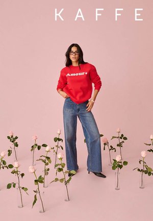 Donna che indossa una felpa rossa con la scritta "Amour" e jeans blu in piedi tra vasi con rose rosa su uno sfondo rosa con la scritta "KAFFE" sopra.