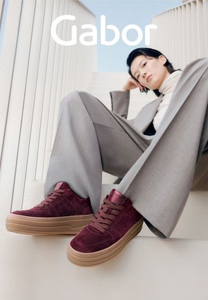 Burgundy suede sneakers with a textured finish, lace-up front, and raised tan rubber sole. Model's gray wide-leg suit complements the footwear.