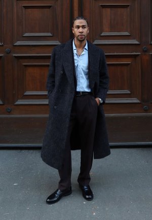 Manteau en laine noire, chemise bleue, pantalon foncé et chaussures noires brillantes. Le manteau possède des boutons croisés et une finition texturée.