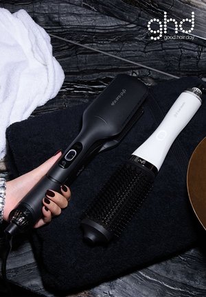 To hårstylingværktøjer på en mørk overflade: en sort varm børste med knapper og en hvid rund børste med sorte børster. Et hvidt håndklæde ligger i nærheden.