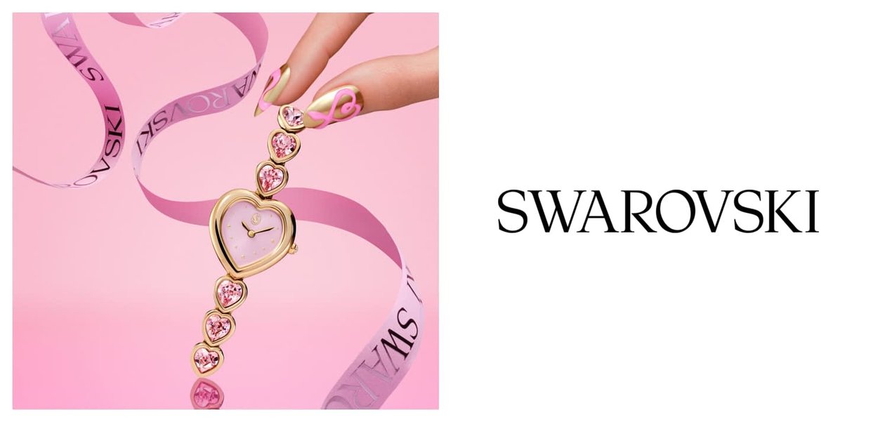 Mano che tiene un orologio a forma di cuore tono oro con cristalli rosa e nastro rosa su sfondo rosa, accanto al logo Swarovski.