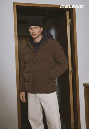 Bruine donzen jas met een capuchon, voorzien van een ritssluiting en zijzakken. Gedragen over een zwarte hoodie, gecombineerd met witte broek. Donkere muts.