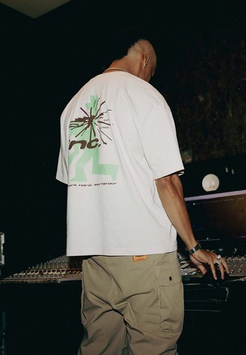 Person mit rasierten Seiten und kurzen Haaren auf dem Oberkopf justiert ein Audio-Mischpult in einem schwach beleuchteten Tonstudio und trägt ein übergroßes T-Shirt mit Grafik.