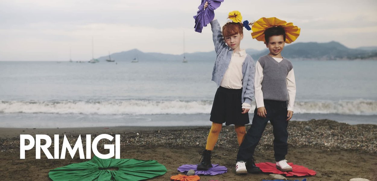 Due bambini stanno su una spiaggia. La ragazza indossa un cardigan azzurro chiaro, una gonna nera e calze gialle alte, mentre tiene un fiore viola; il ragazzo indossa un gilet grigio e pantaloni scuri, con un cappello a forma di fiore giallo. Forme circolari colorate si trovano sulla sabbia.