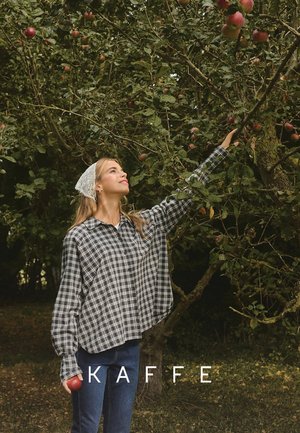 Oversized, grijs geruit shirt van zachte stof met lange mouwen, gecombineerd met blauwe jeans. De achtergrond bevat appelbomen en rode appels.
