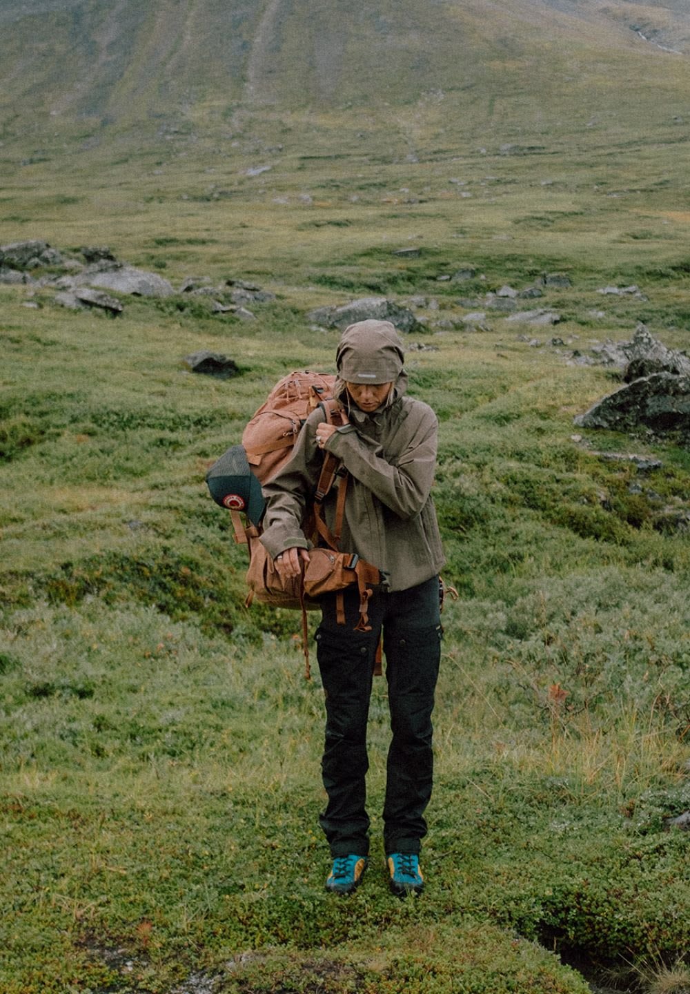 Person som bär en beige jacka och svarta byxor, bär en brun ryggsäck, står på grön gräs med bergig terräng i bakgrunden.