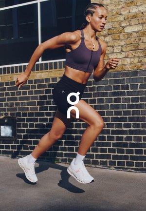 Athletic woman in a dark sports bra and black shorts, running in white shoes with red accents, against a brick wall background.