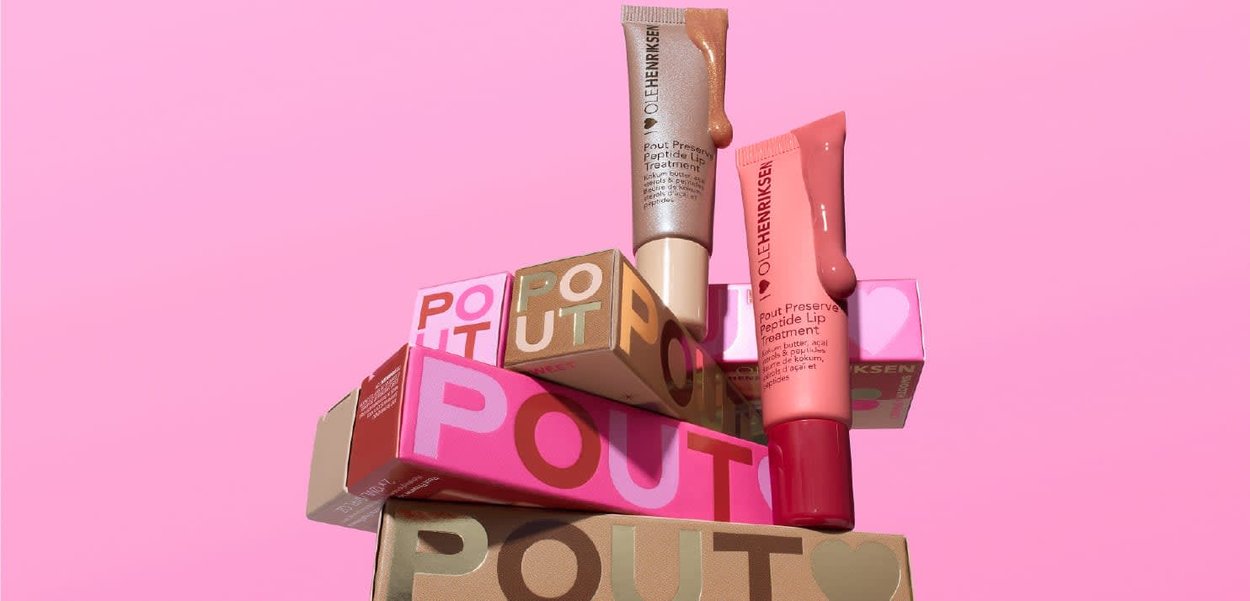 Due tubetti di trattamento per le labbra OLEHENRIKSEN con campioni del prodotto sopra una pila di scatole colorate etichettate "POUT" su uno sfondo rosa.