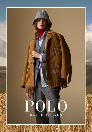 Une jeune femme porte un chapeau seau gris, un col roulé rouge, un costume gris et une veste en daim frangée marron devant un fond beige.