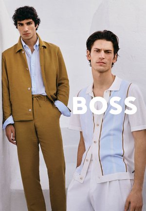 Twee mannen poseren in stijlvolle outfits, één in een bruin jasje en broek, de ander in een wit hemd met blauwe strepen, tegen een witte achtergrond.