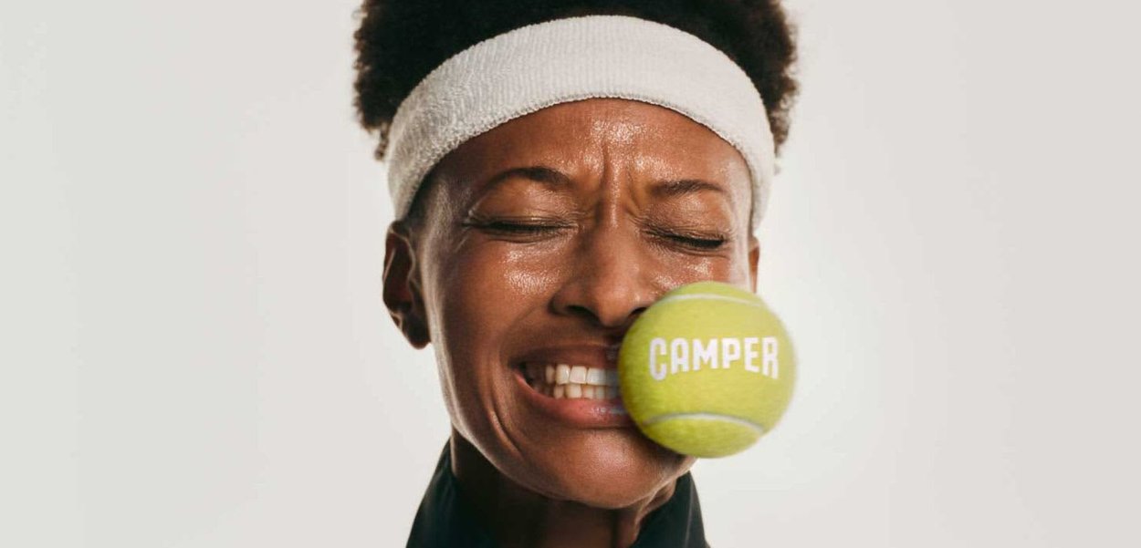 Joven atleta con una diadema blanca reacciona mientras una pelota de tenis con el texto "CAMPER" golpea su mejilla, cerrando los ojos de sorpresa.