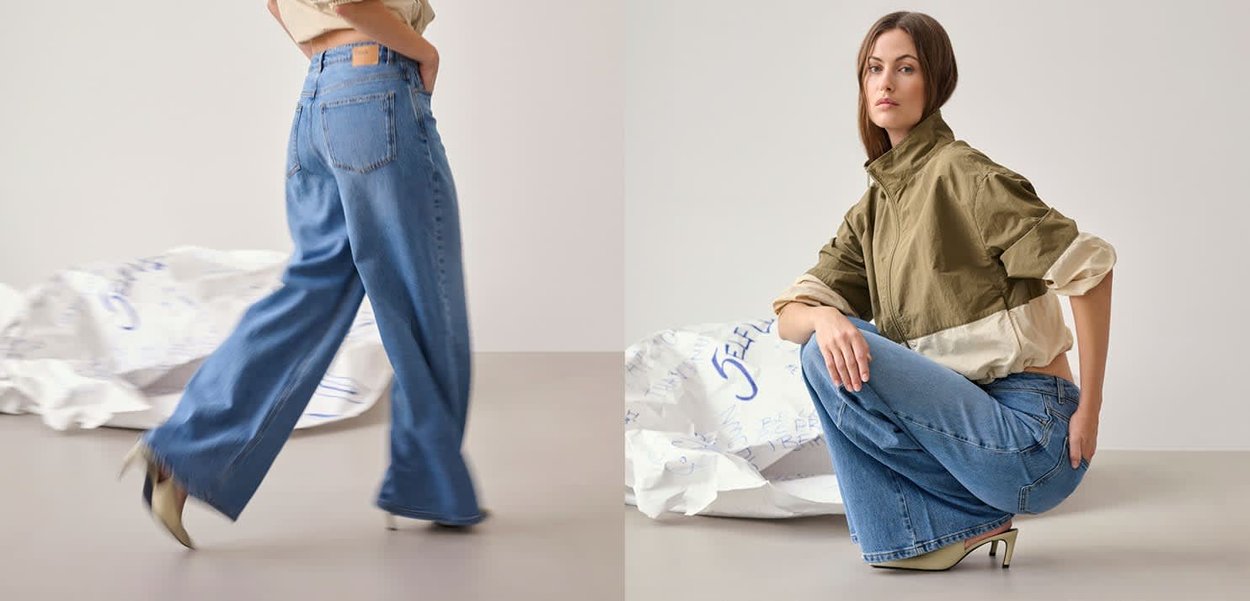 Frau trägt blaue Jeans mit weitem Bein und eine zweifarbige Jacke in Grün und Beige, steht und hockt vor einem neutralen Hintergrund.